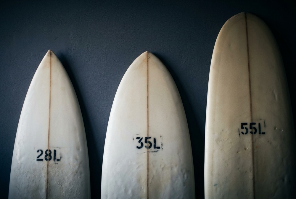 Trois planches de surf debout contre un mur sombre, leur volume peint au pochoir sur le pont : 28L, 35L, 55L.
